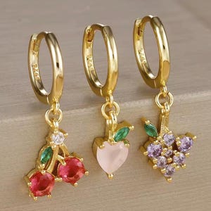 Könnte beinhalten: Drei goldfarbene Creolen mit Anhängern zum Thema Obst. Ein Ohrring hat rote Kirschanhänger, ein anderer einen herzförmigen Pfirsich in Rosa und der dritte eine Traube aus lila Trauben. Jeder Anhänger hat grüne Blattakzente und kleine klare Steine.
