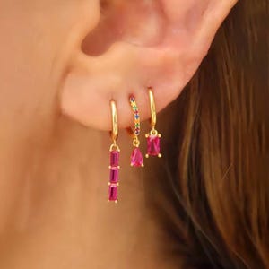 Könnte beinhalten: Drei goldene Ohrringe werden gezeigt. Ein Ohrring hat eine vertikale Reihe von drei rosa rechteckigen Steinen. Ein anderer hat einen kleinen rosa Tropfenstein. Der dritte Ohrring hat einen rosa rechteckigen Stein. Ein dritter Ohrring ist ein goldener Reifen mit bunten Steinen.