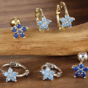Könnte beinhalten: Eine Auswahl an Ohrringen mit Blumenmotiven. Einige haben eine goldene Oberfläche mit blauen Edelsteinen, andere sind silberfarben mit hellblauen Edelsteinen. Die Ohrringe werden auf einer Holzoberfläche präsentiert.