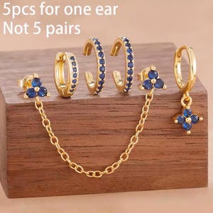 Peut inclure: Un ensemble de cinq boucles d'oreilles dorées avec des pierres bleues, comprenant deux petits anneaux, deux manchettes ouvertes et un anneau avec une breloque fleur suspendue par une chaîne. Texte : 5pcs for one ear Not 5 pairs.