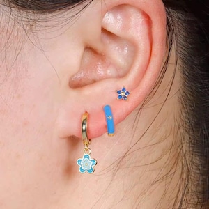 Könnte beinhalten: Nahaufnahme eines Ohrs mit drei Ohrringen. Ein goldener Creolen-Ohrring mit einem hellblauen Blumenanhänger, ein kleiner blauer Creolen-Ohrring und ein blauer Blumenstecker. Die Ohrringe haben blaue und goldene Akzente.