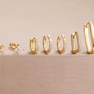 Könnte beinhalten: Eine Auswahl an goldfarbenen Ohrringen, präsentiert auf einer hellbraunen Oberfläche. Die Ohrringe umfassen Stecker und Creolen, einige mit kleinen, klaren Edelsteinen. Die Ohrringe sind in einer Reihe angeordnet und zeigen verschiedene Designs.