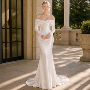 May include: A white, off-the-shoulder wedding gown with long sleeves. The dress features a fitted bodice and a flowing mermaid skirt. The bride is standing on a stone patio with a column and a potted plant in the background.