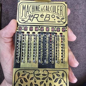 Peut inclure: Ancienne calculatrice mécanique en laiton et noire, avec le texte "MACHINE À CALCULER R e B". Elle comporte plusieurs rangées de cadrans numériques et des motifs floraux décoratifs.