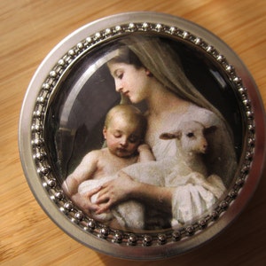 May include: A round, silver-toned metal compact mirror. The mirror's face features a classical painting of a woman, a baby, and a lamb. The edge of the mirror is decorated with small, silver-colored beads.