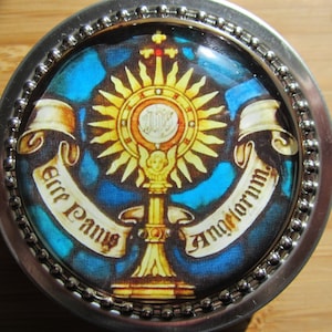 May include: A silver round box with a glass top featuring a religious image of a golden chalice with a host inside, surrounded by a sunburst and two scrolls with the text "Ecce Panis" and "Angelorum".