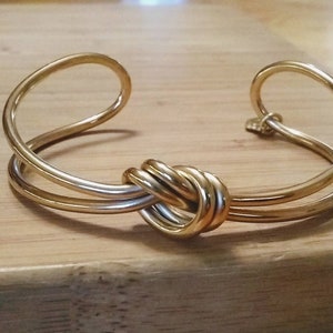 May include: A gold-toned bangle bracelet with a knotted design. The bracelet is made of two bands of metal that are intertwined.