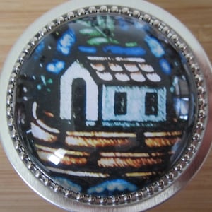 May include: A silver round trinket box with a glass top featuring a colorful stained glass design of a house with a blue roof and windows.