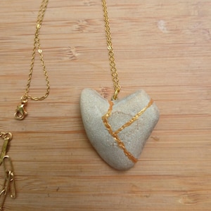 May include: A heart-shaped pendant necklace. The pendant is a light gray stone with gold-colored lines. The necklace chain is gold-colored. The necklace is displayed on a wooden surface.