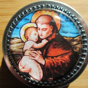 May include: A round metal trinket box with a colorful image of Saint Anthony of Padua holding a baby Jesus. The image is surrounded by a silver beaded border.