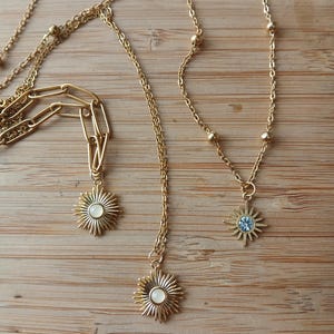 May include: Gold-toned necklaces with sunburst pendants. One necklace features a blue gemstone in the center of the sunburst. The necklaces are displayed on a wooden surface. The chains have different styles, including beaded and paperclip links.
