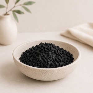 May include: A beige speckled ceramic bowl filled with small, dark charcoal pieces. The bowl is centered in the frame, with a neutral background and a vase with greenery visible in the background.
