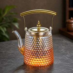 Peut inclure: Une théière en verre transparent avec un motif en nid d'abeille texturé qui passe du transparent en haut à un orange ambré chaud à la base. Elle comprend un couvercle en métal doré avec un fleuron décoratif et une anse dorée arquée.