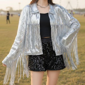 May include: Silver sequin jacket with fringe detail. The jacket has a collared neckline and long sleeves. The fringe hangs from the sleeves and the bottom hem. The jacket is paired with black sequin shorts.
