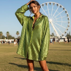 Puede incluir: Una camisa abotonada de manga larga en un vibrante color verde, cubierta de lentejuelas. La camisa tiene un cuello clásico y se lleva abierta, revelando un vistazo del escote del usuario. El fondo presenta una noria y un campo de hierba.