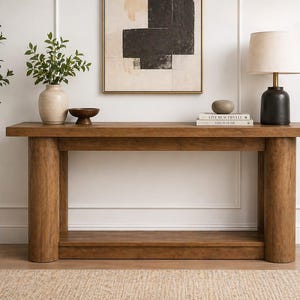 May include: A solid wood console table with a natural finish. The table has a rectangular top and two cylindrical legs on each side. A lower shelf provides additional storage. The table is decorated with a lamp, vase, bowl, and books.