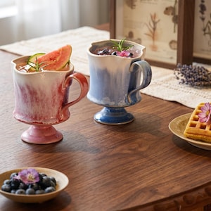 Puede incluir: Dos tazas de cerámica con pedestal y bordes ondulados, una con acabado degradado en rosa y blanco y la otra en azul y blanco. Cada taza presenta un asa curva y una base firme, conteniendo una bebida decorada con trozos de fruta fresca.