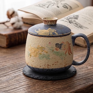 May include: A ceramic mug with a lid, featuring a goldfish design. The mug has a blue handle and lid, and sits on a black coaster. The mug's body is beige with gold and red fish illustrations.