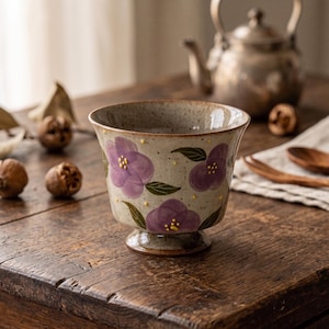 May include: A ceramic footed bowl with a speckled beige finish, adorned with hand-painted purple flowers and green leaves. The bowl has a flared rim and is set on a wooden table, with a teapot in the background.