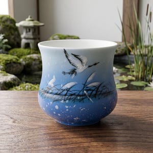 May include: A ceramic cup featuring a gradient blue finish that transitions from a light sky blue at the top to a deeper blue at the base. The exterior is decorated with a hand-painted white crane in flight above dark reeds and white speckles.