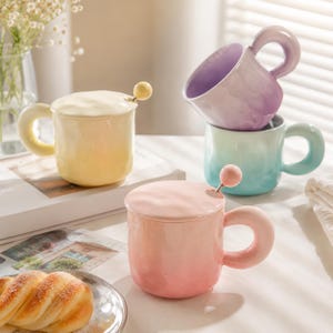 Könnte beinhalten: Eine Sammlung farbenfroher Keramikbecher mit passenden Deckeln und Löffeln. Die Becher sind in Gelb-, Rosa- und Blautönen gehalten und haben abgerundete Griffe. Die Löffel haben eine kleine Kugel am Ende. Die Becher sind auf einer weißen Oberfläche gestapelt.