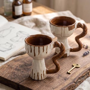 May include: Two ceramic mugs with a cream-coloured base and brown painted details. One mug features vertical brown drips, while the other has a brown grid pattern. Both mugs have a tall, flared base and a distinctively wavy, twisted brown handle.