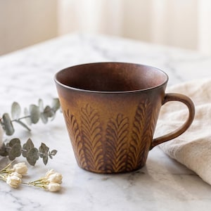 Peut inclure: Une tasse en céramique marron avec un motif texturé en forme de feuilles. La tasse a une anse arrondie et une forme légèrement évasée. L'intérieur est brun foncé et la tasse est posée sur une surface en marbre.
