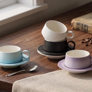 May include: A collection of modern ceramic teacups and saucers in various pastel colors: light blue, black, white, and lavender. A silver spoon rests near the blue cup. The cups are on a wooden surface, with a book and coffee beans in the background.