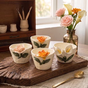 May include: Four off-white ceramic bowls with floral designs. Each bowl features a different flower: a peach rose, an orange rose, an orange lotus, and two white calla lilies. The bowls sit on a wooden board with a gold spoon.