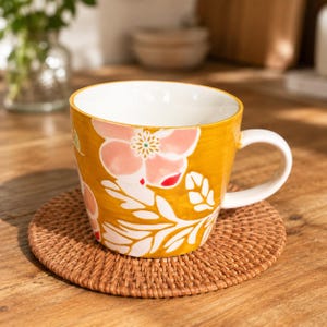 May include: A ceramic mug with a mustard yellow exterior featuring a white and pink floral pattern. The mug has a white interior and a white handle, and it is resting on a round, woven brown rattan coaster. The design includes stylized leaves.