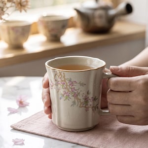 May include: A beige ceramic mug with a floral design, filled with tea, is held over a pink placemat. The mug has a gold rim and a handle. Other teacups and a teapot are visible in the background. The mug is approximately 4 inches tall.