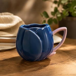May include: A blue ceramic mug in the shape of a tulip flower, featuring a light pink handle. The mug is placed on a wooden surface, with a soft background. This mug is a unique and decorative item for drinks.