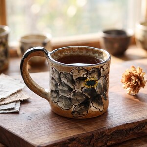 Pode incluir: Uma caneca de cerâmica com um design floral. A caneca está cheia de um líquido escuro, provavelmente chá ou café. O design floral apresenta pétalas pretas e cinzentas com um centro amarelo. A caneca tem uma pega castanha e está sobre uma superfície de madeira.