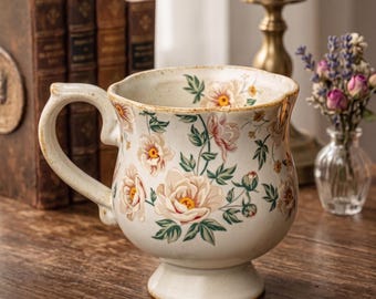 May include: Cream-colored ceramic mug with a pedestal base and a curved handle. The mug is adorned with a floral pattern featuring pink and cream-colored flowers with green leaves. The rim has a slightly distressed, aged look.
