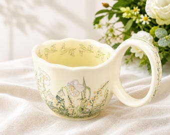 May include: Cream-coloured ceramic teacup with a large, rounded handle. The cup features a floral design in soft green and yellow hues, with a scalloped rim. The handle is also decorated with floral patterns.