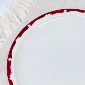 Op de afbeelding: Een wit keramiek bord met een dieprode rand en een decoratieve rand. Het bord heeft een eenvoudig ontwerp met een gegolfde rand.