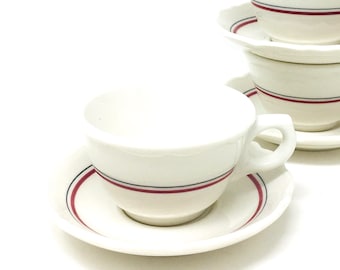 May include: A stack of five white ceramic teacups and saucers. Each cup and saucer features a red stripe design. The cups have a handle and a slightly scalloped rim. The saucers have a matching red stripe.