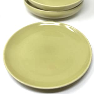 May include: Three round ceramic plates in a muted sage green color. Each plate features a smooth, glossy finish with a subtle, faint geometric grid pattern etched across the surface. The plates are stacked with one in the foreground and two behind.