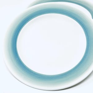 May include: Two white ceramic plates with a gradient blue border. The blue color transitions from a darker shade at the edge to a lighter shade towards the center. The plates are set against a white background.