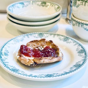 Może przedstawiać: Pojedyncza porcja budyniu chlebowego polana bitą śmietaną i obficie posypana czerwonym dżemem owocowym. Budyń znajduje się na białym talerzu z zielonym wzorem kwiatowym.