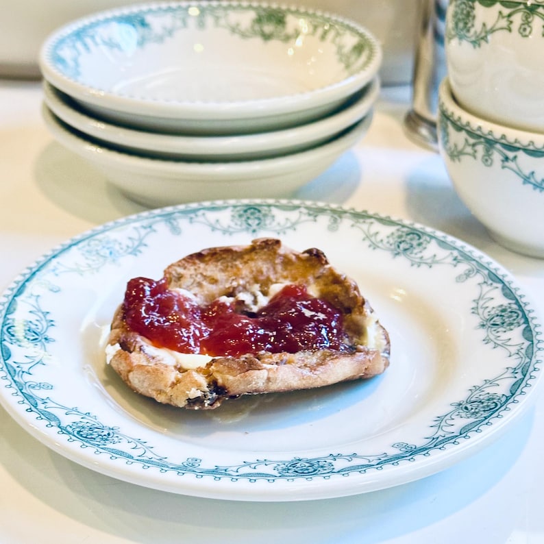 Może przedstawiać: Pojedyncza porcja budyniu chlebowego polana bitą śmietaną i obficie posypana czerwonym dżemem owocowym. Budyń znajduje się na białym talerzu z zielonym wzorem kwiatowym.