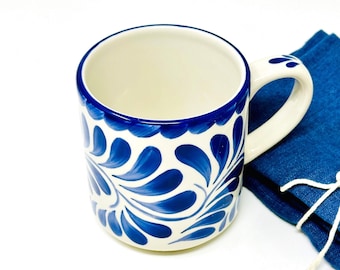May include: A ceramic mug with a white interior and a white handle. The exterior features a blue floral design on a white background. A blue napkin is next to the mug.