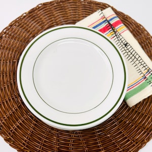 May include: A white plate with a green border sits on a brown woven placemat. A folded cream-colored napkin with colorful stripes and black stitching is next to the plate. The items are arranged on a white surface.