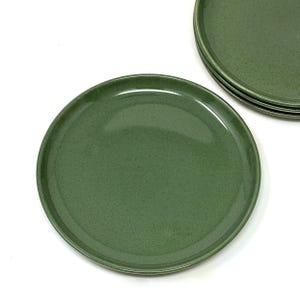 May include: A set of four green ceramic plates. The plates have a speckled texture and a glossy finish. The plates are round and appear to be of a standard size, suitable for serving meals. The plates are arranged on a white surface.