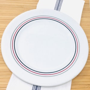 May include: A white ceramic plate with a red, black, and gray striped border. The plate is resting on a white cloth with a blue and gray striped design. The plate is round and has a simple, elegant design.