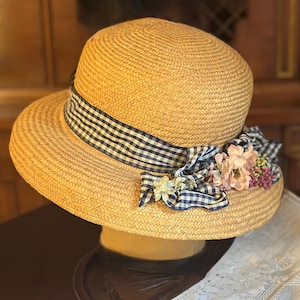 May include: A tan straw hat featuring a rounded crown and a wide brim. A navy blue and white gingham ribbon is wrapped around the base of the crown, tied into a bow on the side. The bow is accented with small, artificial pink and white flowers.