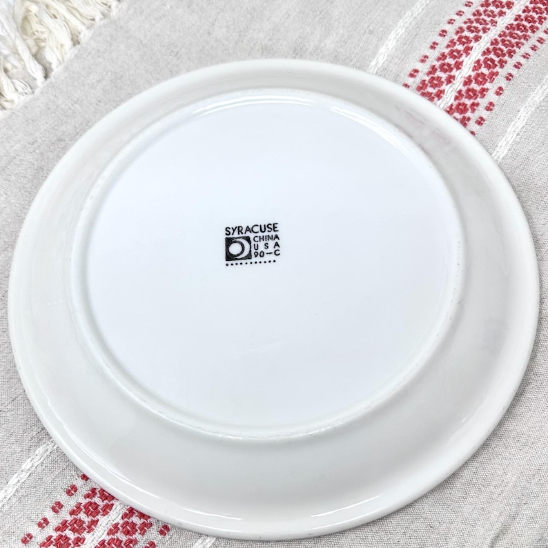 Op de afbeelding: Witte keramische plaat met een zwarte stempel aan de onderkant met de tekst "Syracuse China USA 90-C".