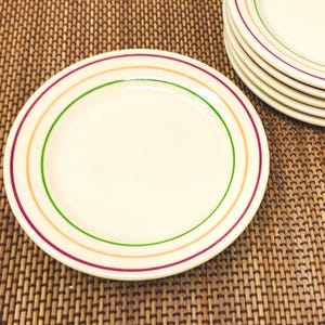 May include: A stack of round, off-white ceramic plates featuring concentric circular bands in green, orange, and dark pink. The plates have a smooth, glossy finish and a simple, clean design, with one plate displayed in the foreground and several others stacked behind it.