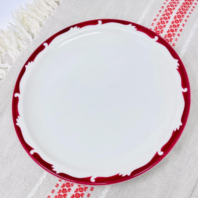 Op de afbeelding: Een wit keramiek bord met een dieprode rand en een decoratieve rand. Het bord heeft een eenvoudig ontwerp met een gegolfde rand.