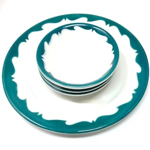 May include: A stack of small white bowls and a large white plate with a teal border and white wave-like accents. The bowls and plate are stacked on top of each other, creating a layered effect. The teal color is a vibrant contrast to the white.